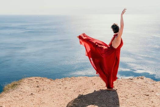 Side view a Young beautiful sensual woman in a red long dress posing on a rock high above the sea during sunrise. Girl on the nature on blue sky background. Fashion photo.