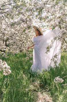 Woman cherry orchard. A happy woman in a long white long dress walks through the green spring blooming cherry garden. Happy cheerful princess bride. The fabric of the skirt flutters in the wind