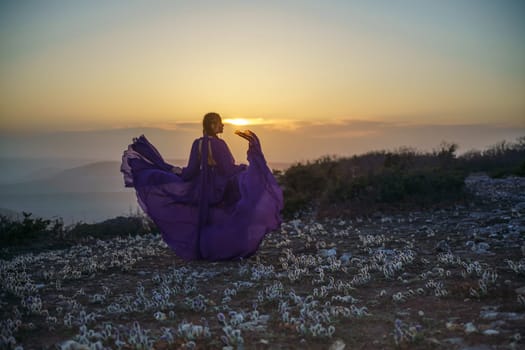 Sunset purple dress woman mountains. Rise of the mystic. sunset over the clouds with a girl in a long purple dress. In the meadow there is a grass dream with purple flowers