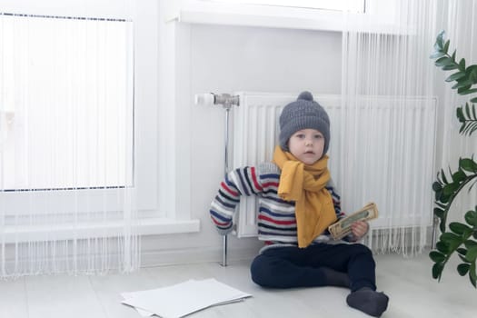 The child is warmly dressed in a sweater and a hat, sitting and holding on to the radiator. The concept of the economic crisis and the lack of heat and heating in homes