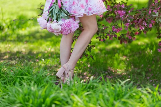 Beautiful legs of a girl in a pink dress, standing with a large bouquet of peonies on green grass, in a blooming garden. Copy space