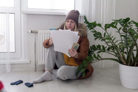 A woman warmly dressed in a brown jacket and hat is sitting near a heating appliance with bills in her hands, money and a phone. The concept of a crisis and a large set for utilities.