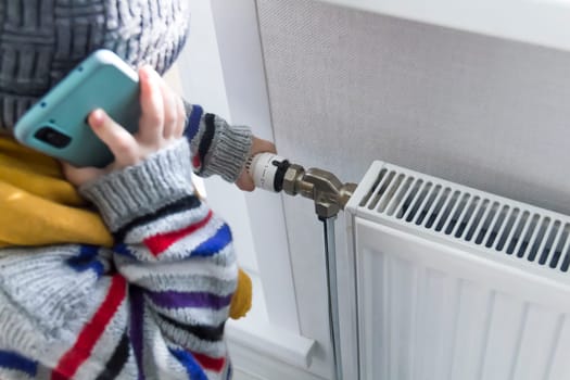 A small child warmly dressed in a sweater and a hat turns the tap on the radiator. The concept of crisis and lack of heating in homes