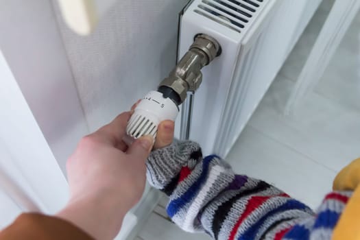 A small child with his mother warmly dressed in a sweater and a hat turns the faucet on the radiator. The concept of crisis and lack of heating in homes