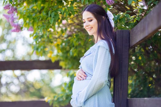a happy pregnant girl with long hair, in a light blue dress stands at the fence, in a lilac garden, on a sunny day. Close up. copy space