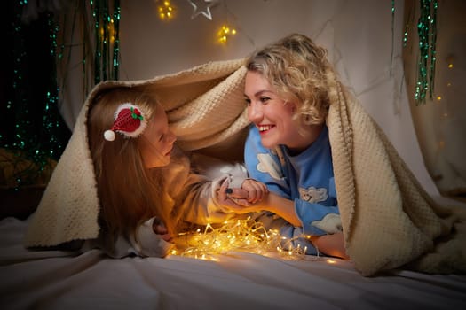 Cute mother and daughter in pajamas lie on soft blanket and having fun in room with Christmas garlands and white background. Tradition of decorating house for holiday. Happy childhood and motherhood