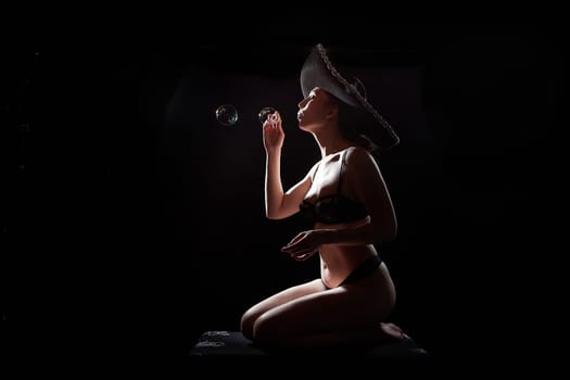 Silhouette of a beautiful young girl with soap bubbles in black underwear and Mexican hat on a dark background. Exotic model posing naked in the studio. Body portrait of sexy girl in usial photoshoot