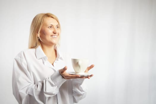 Portrait of pretty blonde smiling woman posing with cup of tea or coffee on white background. Happy girl model in white shirt in studio. The concept of pleasant morning at home or work. Copy space