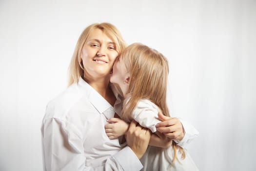 Close portrait of a blonde mother and daughter with hugs on white background. Mom and little girl models pose in the studio. The concept of love, friendship, caring in the family