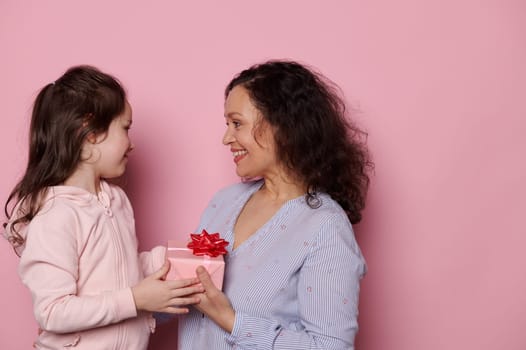 Dark-haired beautiful middle-aged Latin American woman, smiling and feeling touched by a cute present from her lovely daughter for Mother's Day. Portrait of a mom and child on isolated pink background