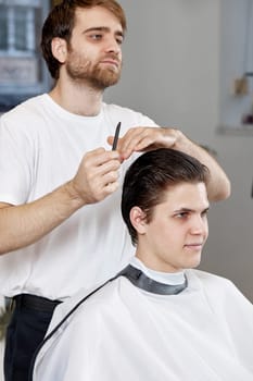 professional male hairstylist combing caucasian man while sitting in chair before haircut at barbershop. client tells what haircut he wants