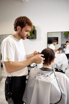 young caucasian man getting haircut by professional male hairstylist using comb and grooming scissors at barber shop.