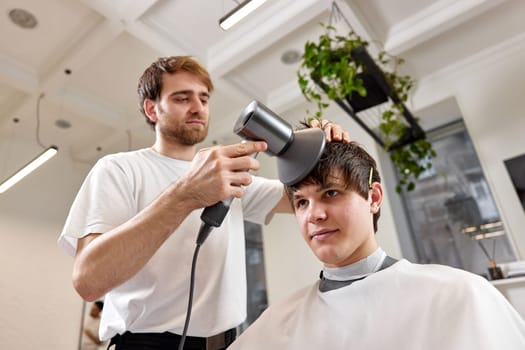 Professional hairdresser using hair dryer and hairbrush of his client in barber shop. Haircut in the barbershop.