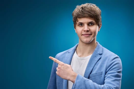 positive caucasian young man in jacket pointing finger at copy space on blue background