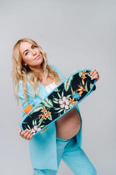 a pregnant girl in turquoise clothes with a skateboard in her hands on a gray background.
