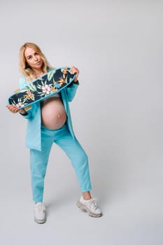 a pregnant girl in turquoise clothes with a skateboard in her hands on a gray background.