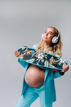 A pregnant girl in a turquoise suit with a skateboard in her hands and headphones stands on a gray background.