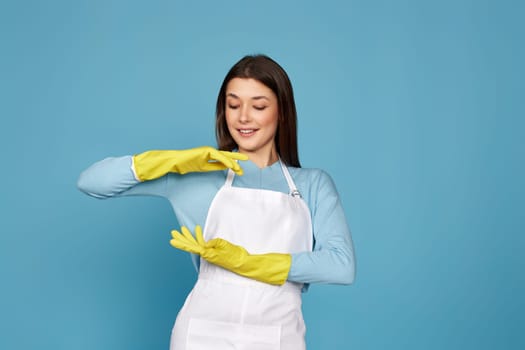 beautiful brunette woman in yellow rubber gloves and cleaner apron showing measure symbol gesture on blue background. cleaning