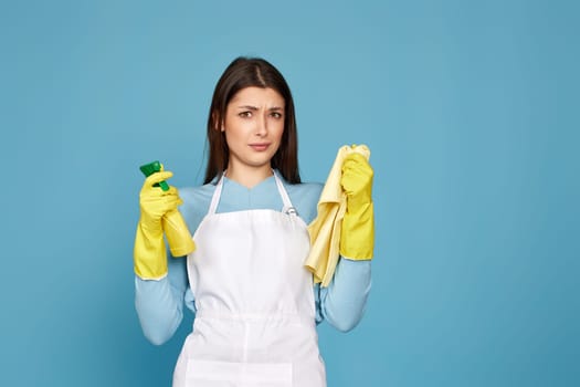 skeptic and nervous girl in gloves and cleaner apron with cleaning rag and detergent sprayer on blue background.