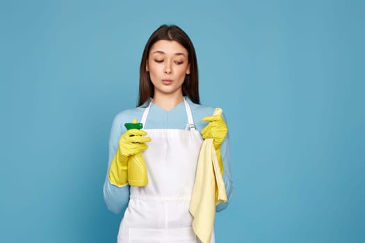 caucasian woman in rubber gloves and cleaner apron with cleaning rag and detergent sprayer on blue background.
