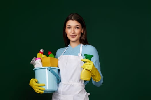 beautiful woman in yellow rubber gloves and cleaner apron holding bucket with cleaning supplies and detergent sprayer on dark green background.