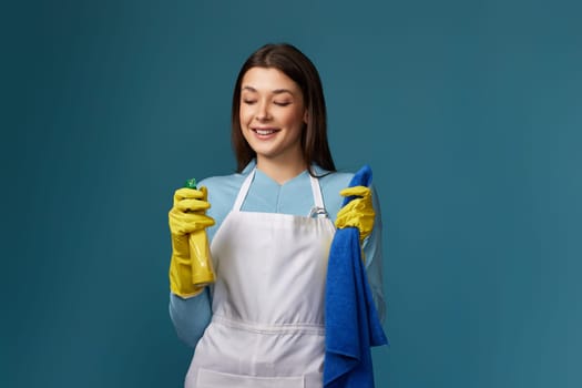 smiling caucasian woman in yellow rubber gloves and cleaner apron with cleaning rag and detergent sprayer on dark blue background.