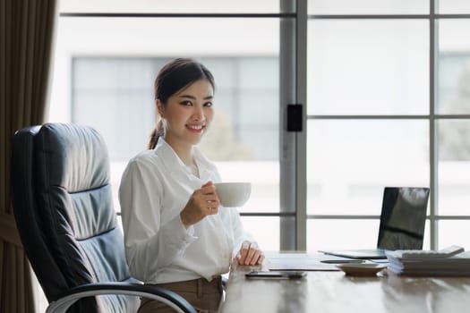 Attractive asian businesswoman sitting and drinking coffee at the desk in home office.