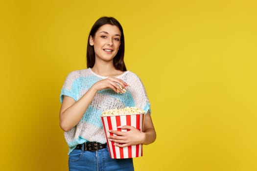 beautiful brunette girl in blue pullover eating popcorn isolated on yellow background