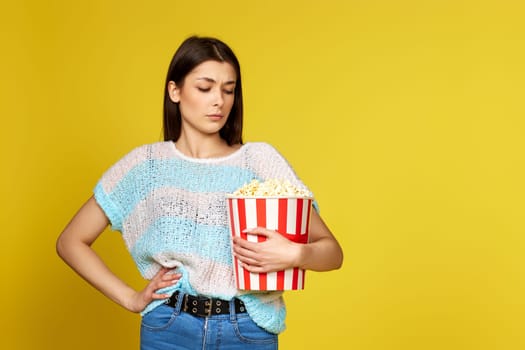 beautiful brunette girl looks doubtfully at popcorn on yellow background. unhealthy food