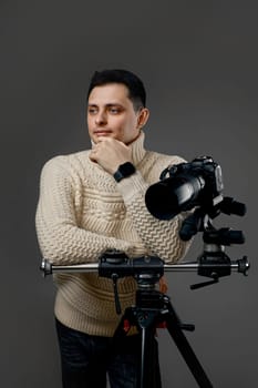 Professional young handsome photographer in sweater near digital camera on tripod on gray background
