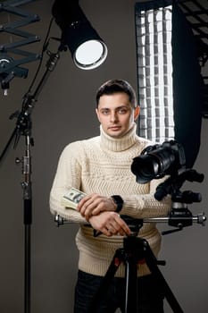 Professional handsome photographer with digital camera on tripod holding hundred dollar bills on gray background with lighting equipment
