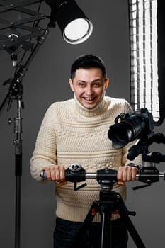 Professional happy photographer with digital camera on tripod on gray background with lighting equipment