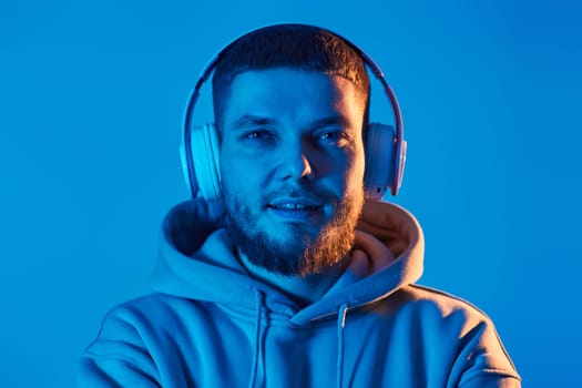 handsome bearded man with headphones in sweatshirt enjoying favorite music on blue neon background. Neon lighting