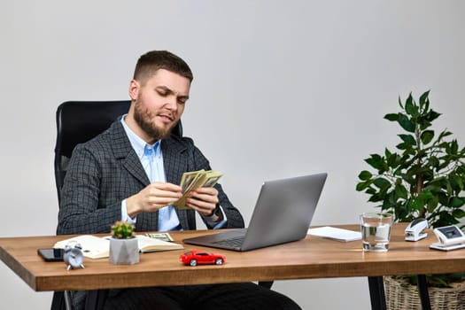 young bearded businessman working on laptop and holding money while sitting on chair at desk.
