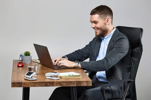 happy bearded businessman typing on computer keyboard, sending emails to business partners, sitting on chair at desk