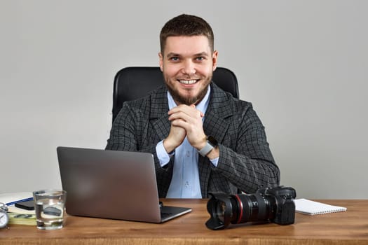 successful bearded male photographer smiling cheerfully while working at desk. dslr camera on table