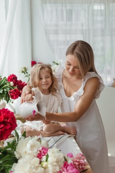 A little blonde girl with her mom on a kitchen countertop decorated with peonies. The concept of the relationship between mother and daughter. Spring atmosphere.