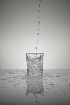 Vertical shot of water pouring into crystal glass with droplets around, on a white shiny table