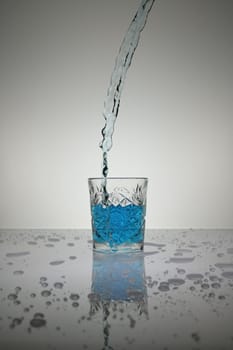 Vertical shot of blue liquid pouring into crystal glass with droplets around,on a white shiny table