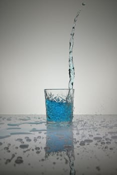 Vertical shot of water pouring into crystal glass with droplets around, on a white shiny table