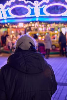 Rear of an old woman with gray hair with a headscarf in front of a colorfully illuminated children's carousel
