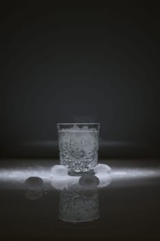 Vertical shot of crystal water glass, ice cubes and waterdrops around, against a dark background
