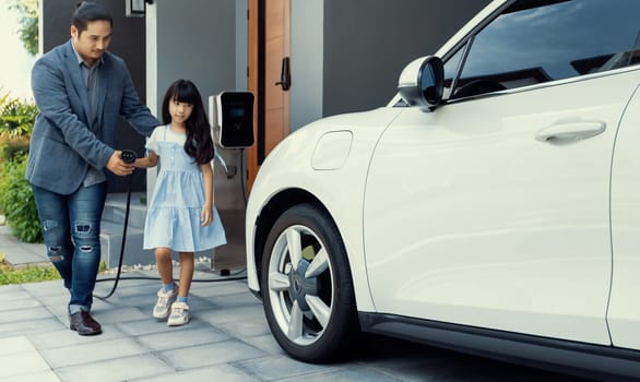 Progressive father and daughter plugs EV charger from home charging station to electric vehicle. Future eco-friendly car with EV cars powered by renewable source of clean energy.