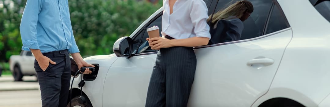 Progressive businessman and businesswoman with coffee, standing at electric car connected to charging station before driving around city center. Eco friendly rechargeable car powered by clean energy.