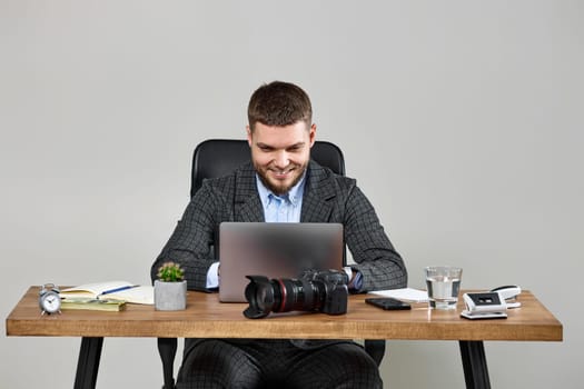 Professional male photographer smiling cheerfully while working on laptop computer at desk. dslr camera on table