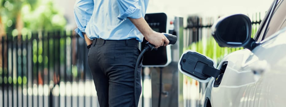 Closeup progressive man holding EV charger plug from public charging station for electric vehicle with background of residential building as concept eco-friendly sustainability energy car concept.