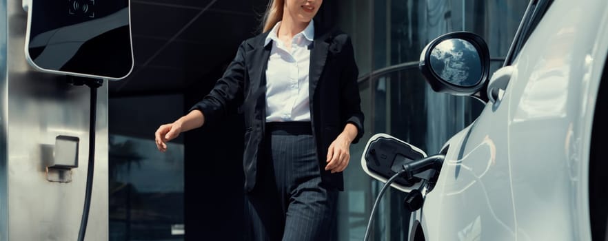Closeup progressive businesswoman wearing suit with electric car recharging at public charging station at modern city center. Eco friendly rechargeable car powered by alternative clean energy.