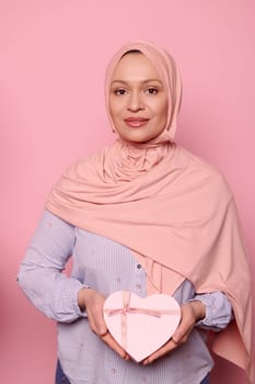 Attractive confident Middle-Eastern Muslim woman in pink hijab, showing at camera a heart shaped gift box on isolated background. Happy Mother's Day. Women's Day. Celebrating festive event, occasion