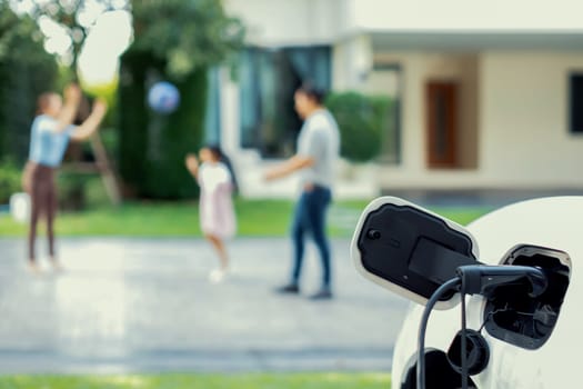 Focus EV charger connected with EV car at outdoor home charging station with blur progressive family playing together in the background. EV car for alternative clean energy concept.