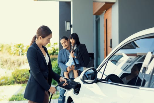 Progressive young parents and daughter with electric vehicle and home charging station. Green and clean energy from electric vehicles for healthy environment. Eco power from renewable source at home.